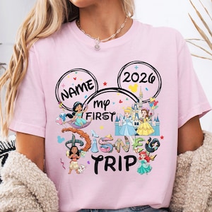 A woman wearing a pink t-shirt with a Disney-themed design, including characters from the Disney film series and the text "Name my first Disney trip".