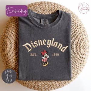 A gray t-shirt with the text "Disneyland EST. 1955" and a Minnie Mouse graphic, placed on a woven basket.