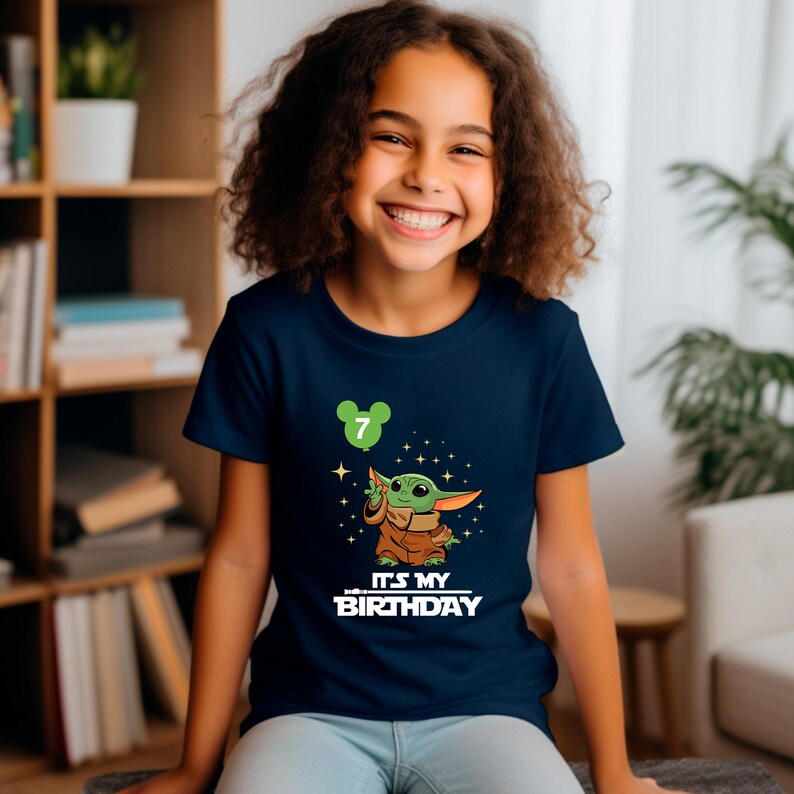 A young girl with curly hair is smiling and wearing a blue t-shirt with a graphic of a character from the Star Wars franchise and the text "It's my birthday".