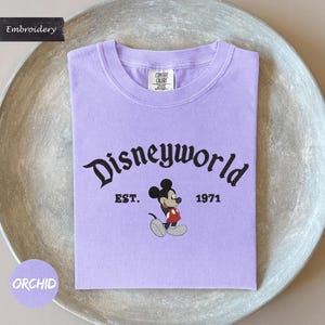 A purple t-shirt with the text "Disneyworld" and a Mickey Mouse graphic, along with the text "EST. 1971" printed on it.