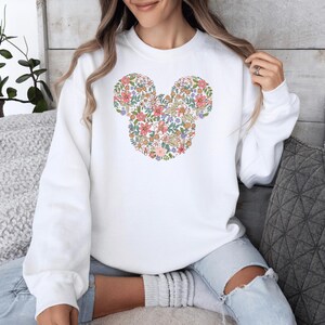 May include: White sweatshirt featuring a floral Mickey Mouse design. The flowers are in shades of pink, yellow, and green.