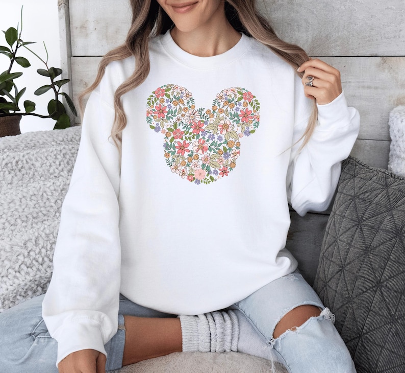 May include: White sweatshirt featuring a floral Mickey Mouse design. The flowers are in shades of pink, yellow, and green.