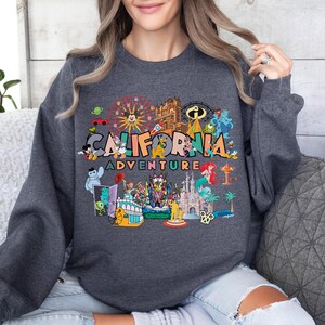 A woman wearing a gray sweatshirt with a colorful graphic design featuring various Disney characters and landmarks, including the Eiffel Tower, Mickey Mouse, and the California Adventure theme park.