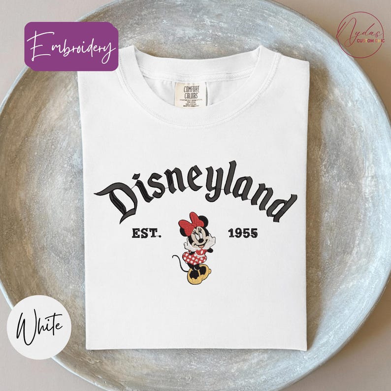 A white t-shirt with the text "Disneyland EST. 1955" and a Minnie Mouse graphic printed on it, placed on a gray plate.
