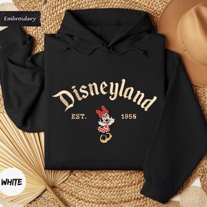 A black hoodie with the word "Disneyland" and the year "1955" printed on it, along with a Minnie Mouse character. The hoodie is laid out on a woven surface, and there is a hat and a fan visible in the background.