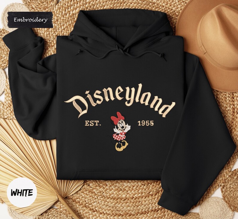 A black hoodie with the word "Disneyland" and the year "1955" printed on it, along with a Minnie Mouse character. The hoodie is laid out on a woven surface, and there is a hat and a fan visible in the background.