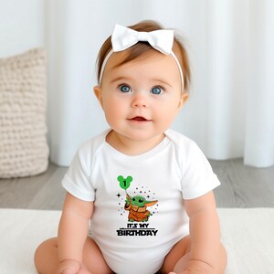 A baby girl wearing a white onesie with a graphic of a character from the Star Wars franchise and the text "It's My Birthday" is sitting on the floor.