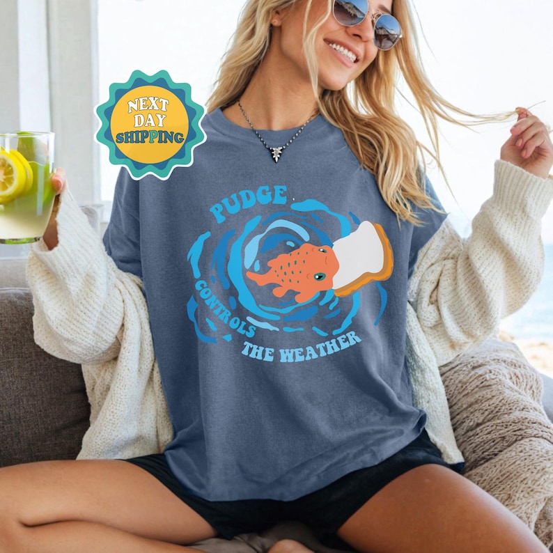 以下が含まれることがあります： 頭の上にパンの切れ端を乗せた魚のグラフィックが描かれた青いTシャツ。Tシャツのテキストは「Pudge Controls The Weather」です。