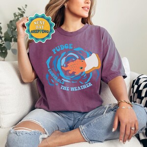 以下が含まれることがあります： 頭の上にパンの切れ端を乗せた魚のグラフィックが描かれた紫色のTシャツ。Tシャツの文字は「Pudge Controls The Weather」です。