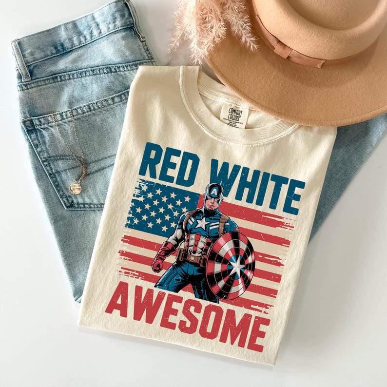 A white t-shirt with a graphic design of a superhero character wearing a red, white, and blue outfit, along with the text "RED WHITE AWESOME" printed on it. The t-shirt is placed on top of a pair of jeans and a beige hat.