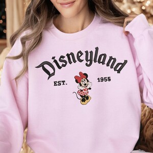 A woman wearing a pink sweatshirt with the text "Disneyland EST. 1955" and a Minnie Mouse graphic.