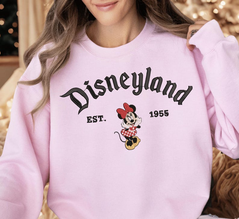 A woman wearing a pink sweatshirt with the text "Disneyland EST. 1955" and a Minnie Mouse graphic.