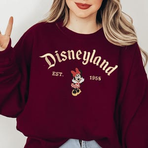 A woman wearing a burgundy sweater with the text "Disneyland EST. 1955" and a Minnie Mouse graphic.