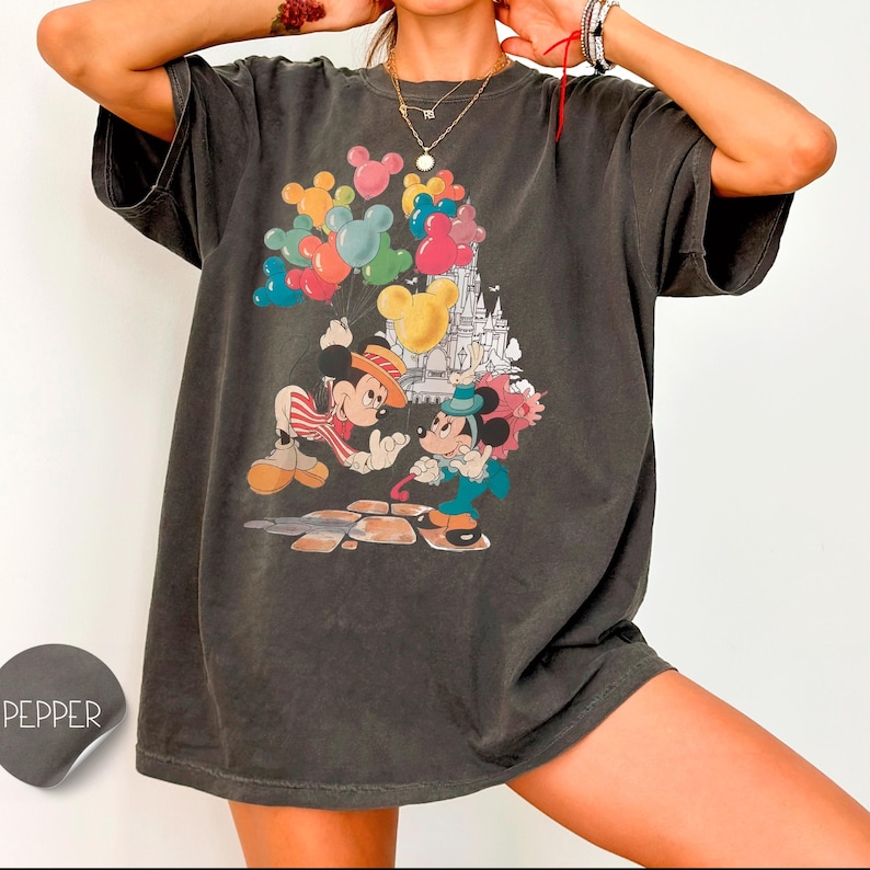 A person wearing a gray t-shirt with a colorful graphic of Mickey Mouse and Minnie Mouse on a castle, along with various other cartoon characters and balloons.