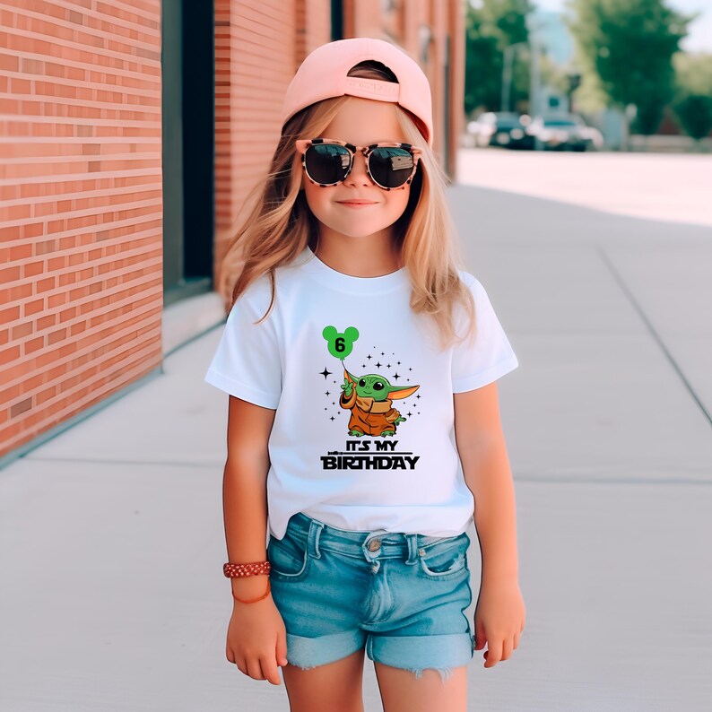 A young girl wearing a white t-shirt with a graphic of a character, sunglasses, and a pink cap is standing on a sidewalk.