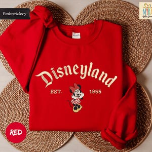 A red Disneyland sweatshirt with the text "Disneyland EST. 1955" and a Minnie Mouse graphic, placed on a woven placemat.