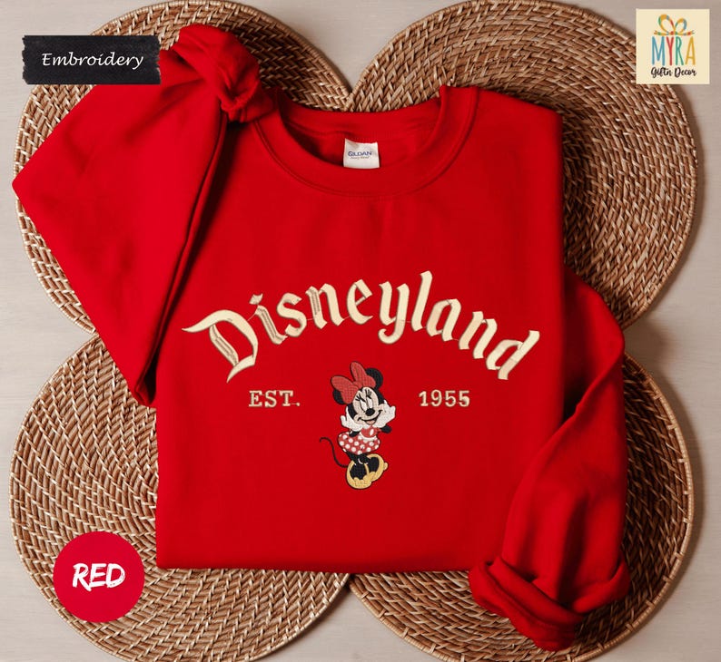 A red Disneyland sweatshirt with the text "Disneyland EST. 1955" and a Minnie Mouse graphic, placed on a woven placemat.