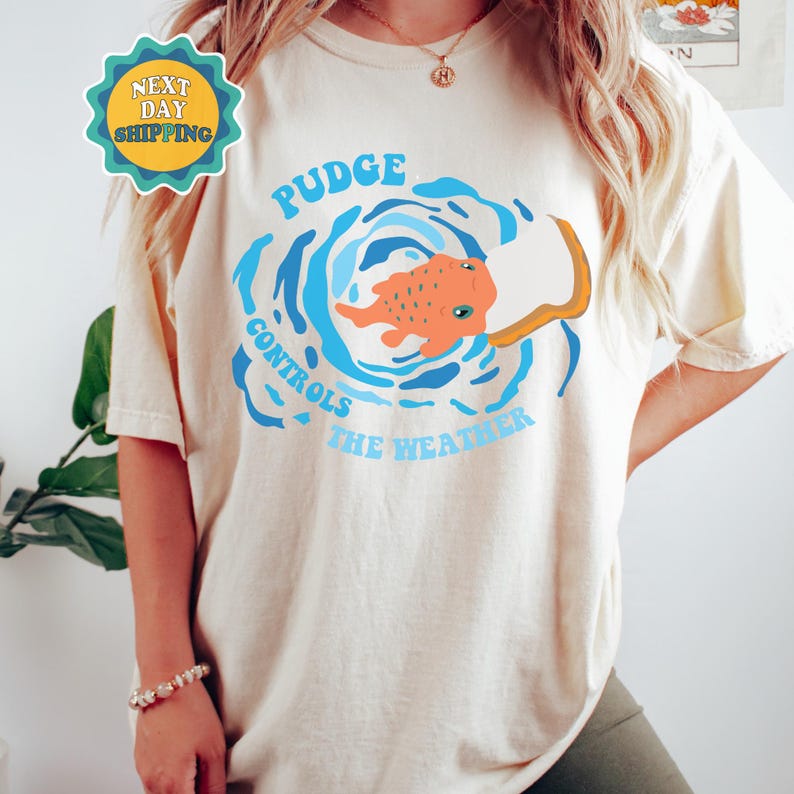 以下が含まれることがあります： 白いTシャツに、青とオレンジの魚のデザインがプリントされています。円の中に「PUDGE CONTROLS THE WEATHER」という文字が書かれています。