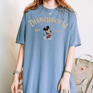 A person wearing a blue t-shirt with the Disneyworld logo and text, standing in front of a beige backpack.