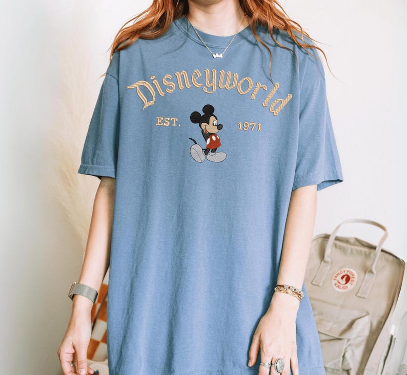 A person wearing a blue t-shirt with the Disneyworld logo and text, standing in front of a beige backpack.