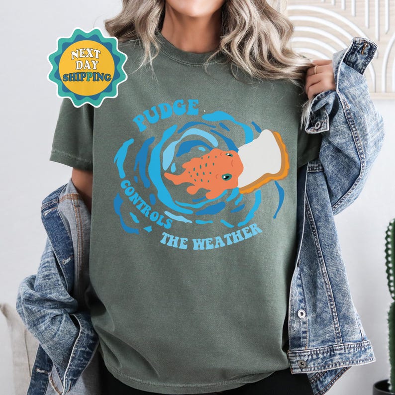 以下が含まれることがあります： 水しぶきの中に魚が描かれた緑色のTシャツ。Tシャツには「PUDGE CONTROLS THE WEATHER」という文字がプリントされています。