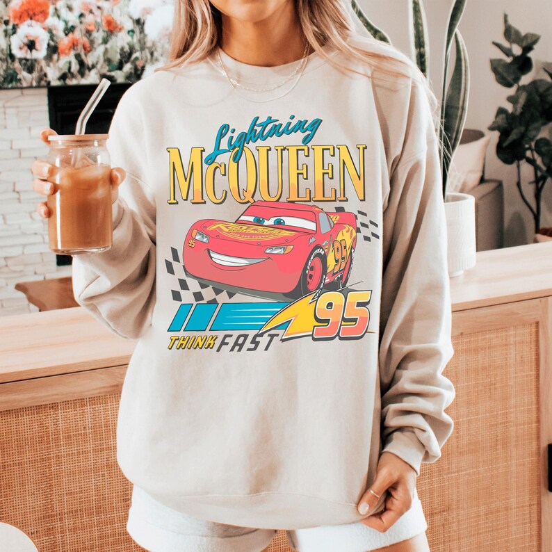 A person wearing a white sweatshirt with a graphic of Lightning McQueen from the Cars movie, holding a drink.