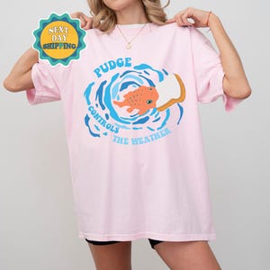 以下が含まれることがあります： 青い渦の中に魚が描かれたピンクのTシャツ。"Pudge Controls The Weather"という文字が書かれている。
