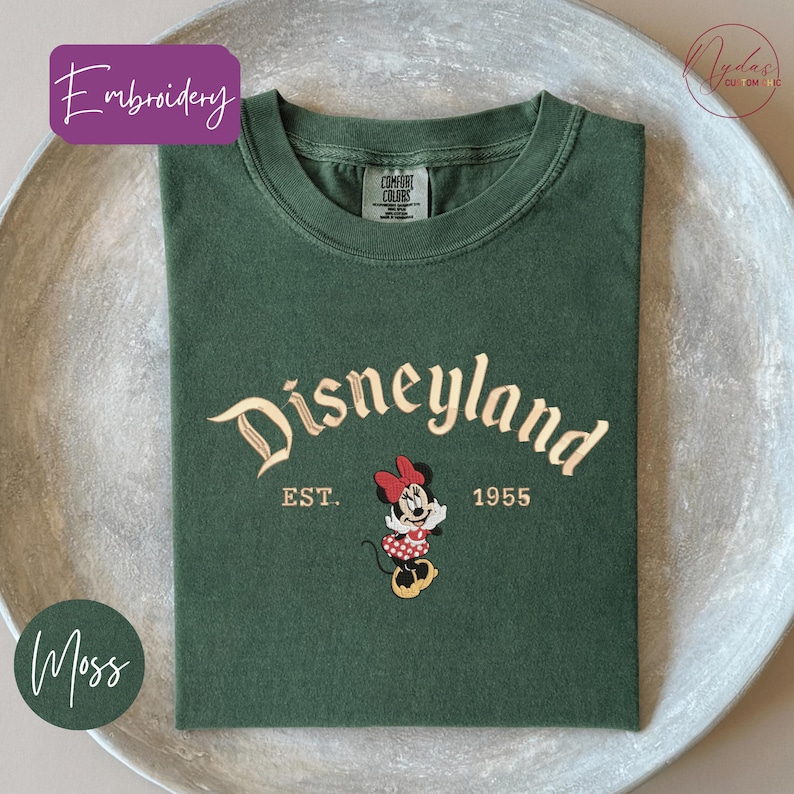 A green t-shirt with the text "Disneyland EST. 1955" and an embroidered Minnie Mouse character, placed on a white surface.