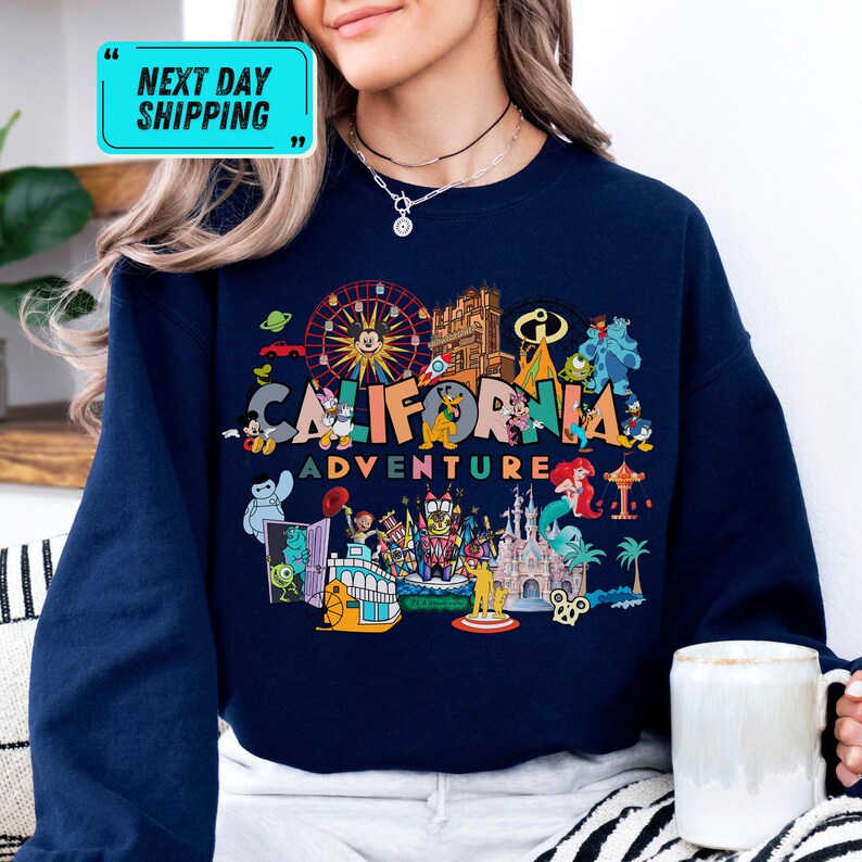 A woman wearing a blue sweatshirt with a colorful graphic design depicting various California-themed elements, such as a ferris wheel, castle, and palm trees.