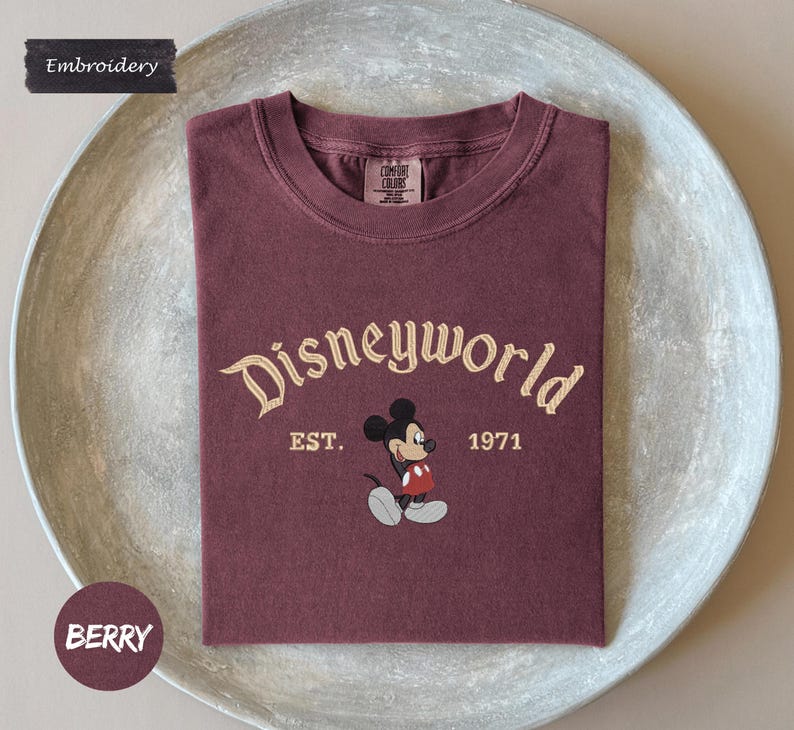 A maroon t-shirt with the text "Disneyworld" and "EST. 1971" printed on it, along with a Mickey Mouse graphic. The t-shirt is displayed on a white plate.