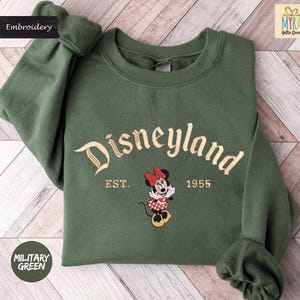 A green sweatshirt with the text "Disneyland EST. 1955" and an embroidered Minnie Mouse character, indicating the establishment year of the theme park.