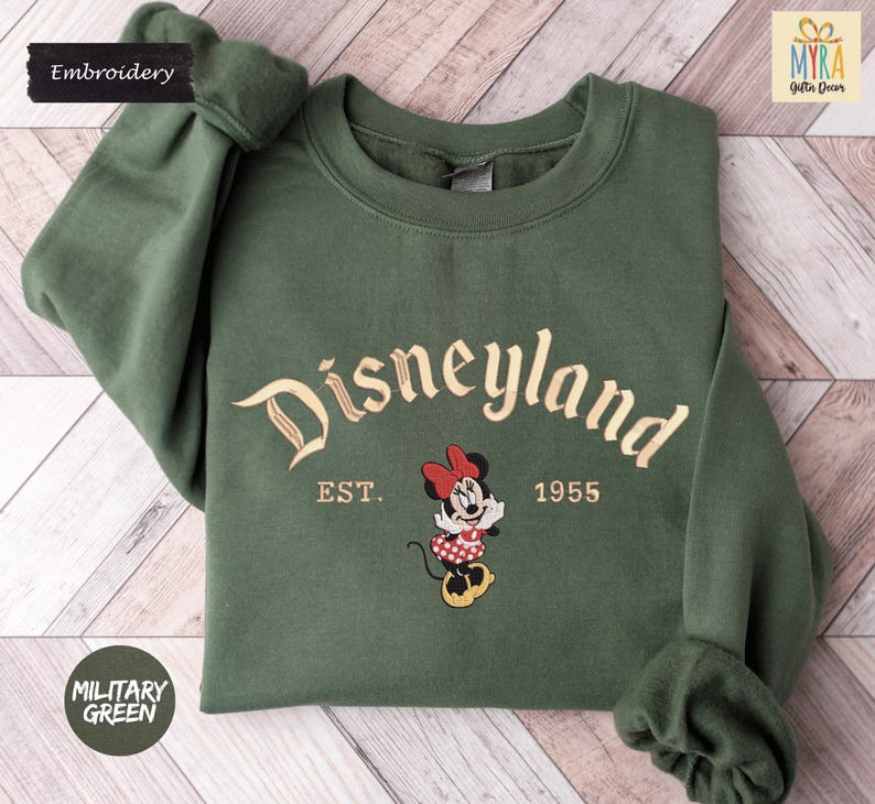 A green sweatshirt with the text "Disneyland EST. 1955" and an embroidered Minnie Mouse character, indicating the establishment year of the theme park.