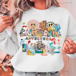 A person wearing a white sweatshirt with a colorful graphic design featuring various Disney characters and landmarks, including a ferris wheel, castle, and palm trees, along with the text "California Adventure".