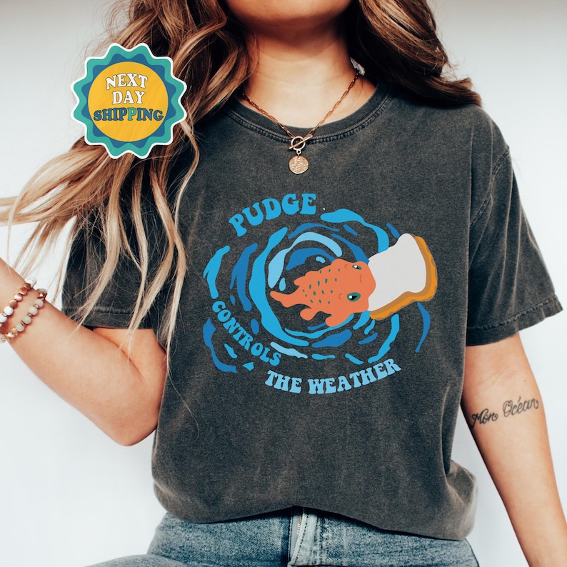 以下が含まれることがあります： パンを食べる漫画の魚のグラフィックがプリントされたダークグレーのTシャツ。魚を囲むように「Pudge Controls The Weather」というテキストがプリントされています。
