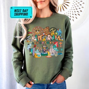 A woman wearing a green sweatshirt with a colorful graphic design featuring various landmarks and icons associated with California, such as the Golden Gate Bridge, Disneyland, and the Hollywood sign.