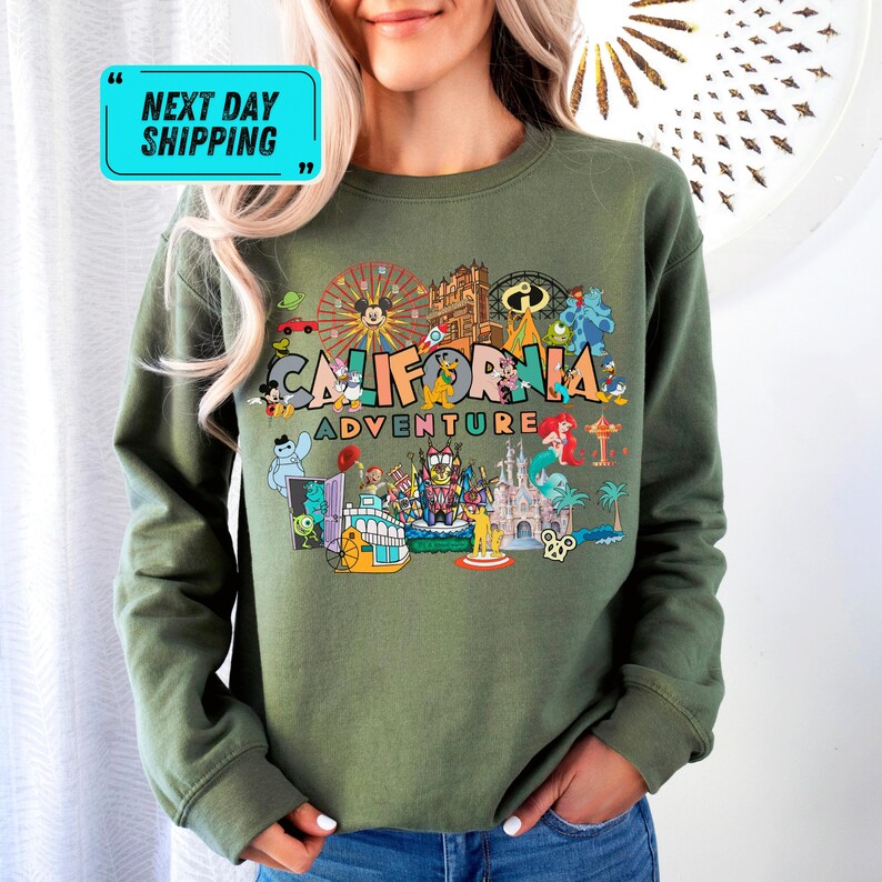 A woman wearing a green sweatshirt with a colorful graphic design featuring various landmarks and icons associated with California, such as the Golden Gate Bridge, Disneyland, and the Hollywood sign.