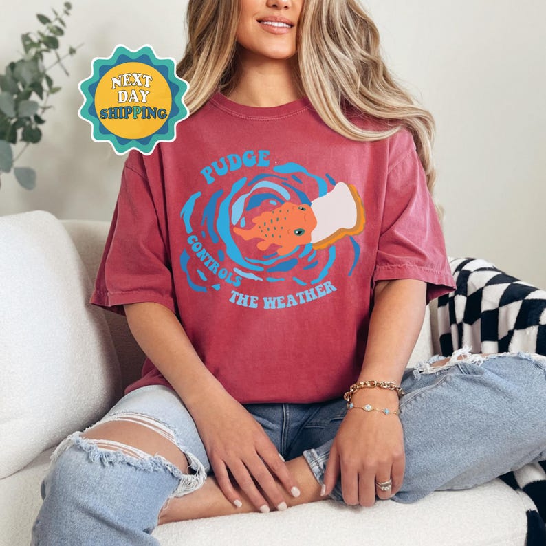以下が含まれることがあります： パンを食べるアニメーションの魚が描かれた赤いTシャツ。Tシャツの文字は「Pudge Controls The Weather」です。