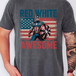 A person wearing a gray t-shirt with a graphic design featuring a superhero character and the text "RED WHITE AWESOME" printed on it.