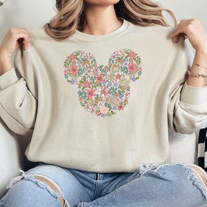May include: A beige sweatshirt featuring a floral Mickey Mouse design. The flowers are in shades of pink, purple, and yellow.