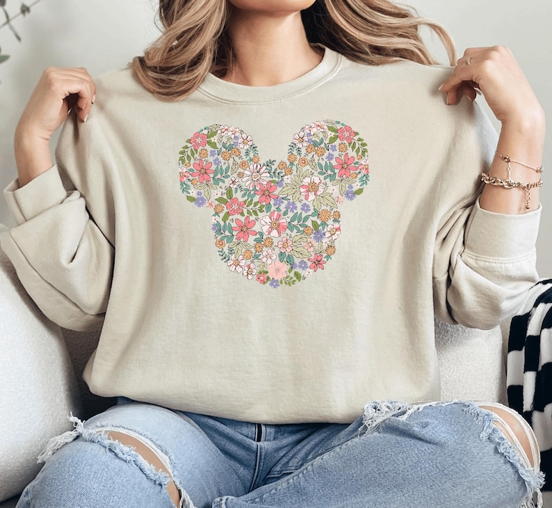 May include: A beige sweatshirt featuring a floral Mickey Mouse design. The flowers are in shades of pink, purple, and yellow.