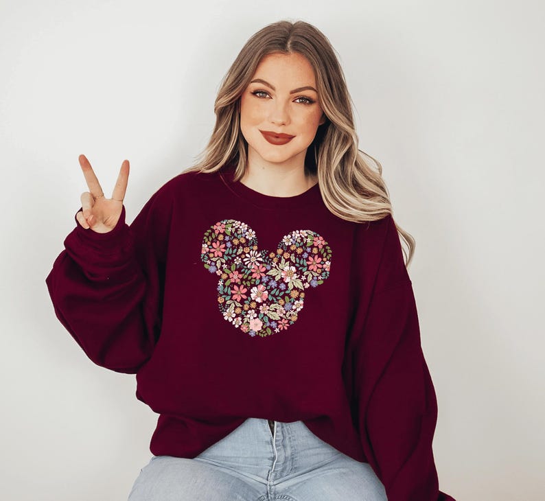 May include: A maroon sweatshirt featuring a floral Mickey Mouse design. The sweatshirt has long sleeves and a crew neck.