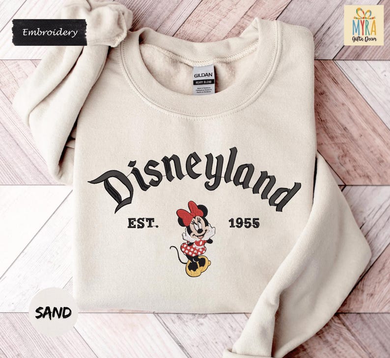 A beige sweater with the text "Disneyland EST. 1955" and a Minnie Mouse graphic printed on it, along with the text "SAND" at the bottom.