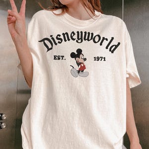 A person wearing a white t-shirt with the text "Disneyworld" and a Mickey Mouse graphic, along with the text "EST. 1971" printed on it.