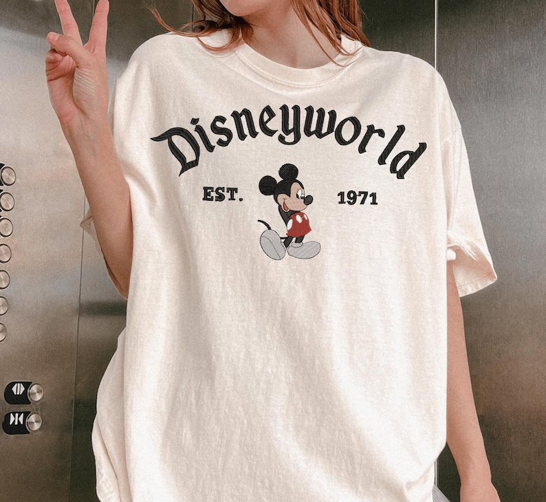 A person wearing a white t-shirt with the text "Disneyworld" and a Mickey Mouse graphic, along with the text "EST. 1971" printed on it.