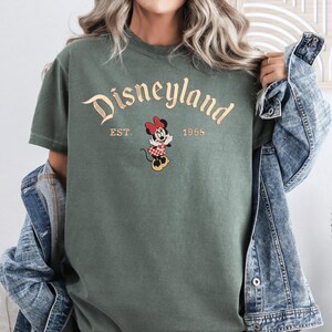 A woman wearing a green t-shirt with the text "Disneyland EST. 1955" and a Minnie Mouse graphic, along with a denim jacket.