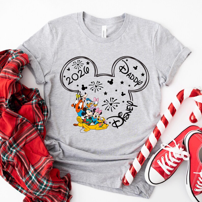 A gray t-shirt with a Disney-themed design, featuring Mickey Mouse and Donald Duck, along with the text "2020 Daddy" and "Disney" printed on it. The t-shirt is placed on a white surface, surrounded by a red plaid scarf, a pair of red and white striped candy canes, and a pair of red and white sneakers.