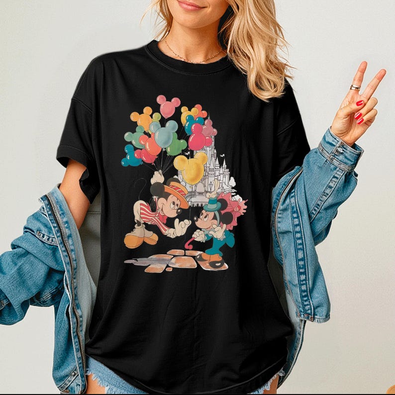 A woman wearing a black t-shirt with a Mickey Mouse and Minnie Mouse design, along with colorful balloons, is posing with her hands in a peace sign.