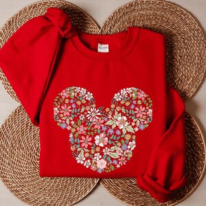 May include: Red sweatshirt featuring a floral Mickey Mouse design. The sweatshirt is folded and placed on a woven surface.
