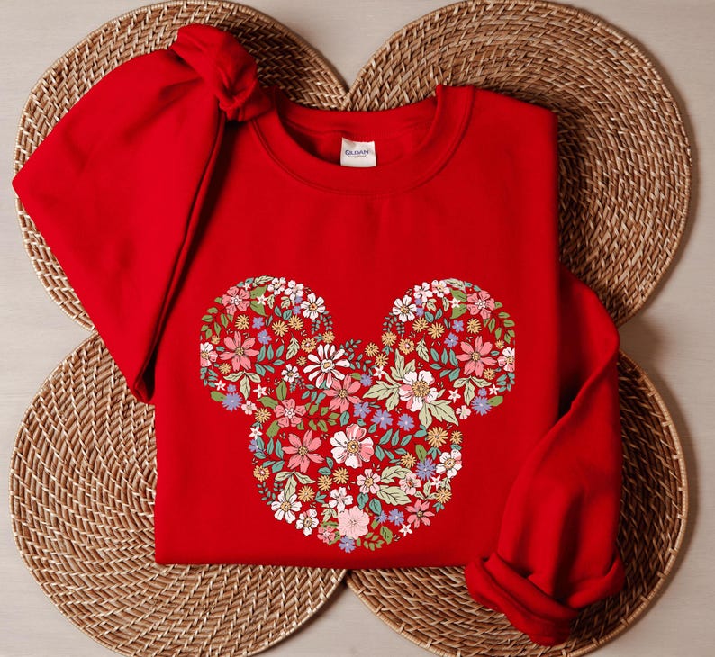 May include: Red sweatshirt featuring a floral Mickey Mouse design. The sweatshirt is folded and placed on a woven surface.