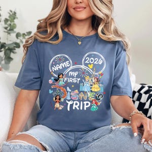 A woman wearing a blue t-shirt with a Disney-themed design, including characters from Disney movies and the text "Name my first trip" printed on it.